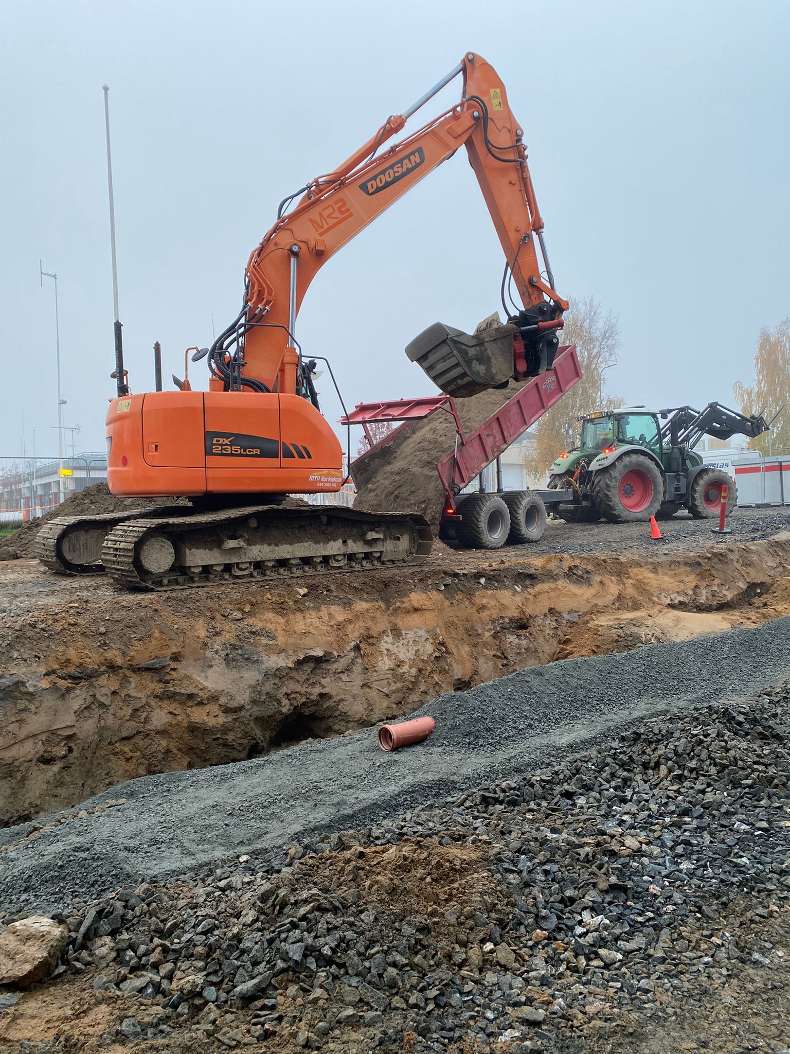 Oranssi Doosan-kaivinkone tyhjentää maata peräkärryyn, vihreä traktori taustalla, harmaa sää.