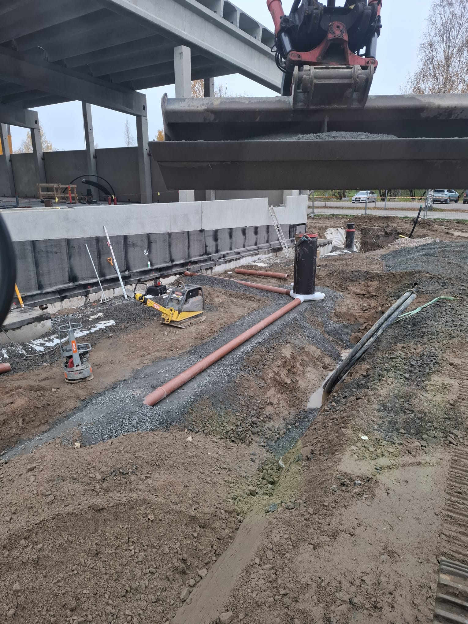 Rakennustyömaa, jossa näkyy putkia, tiivistyskoneita ja kaivinkoneen kauha, harmaa sää.