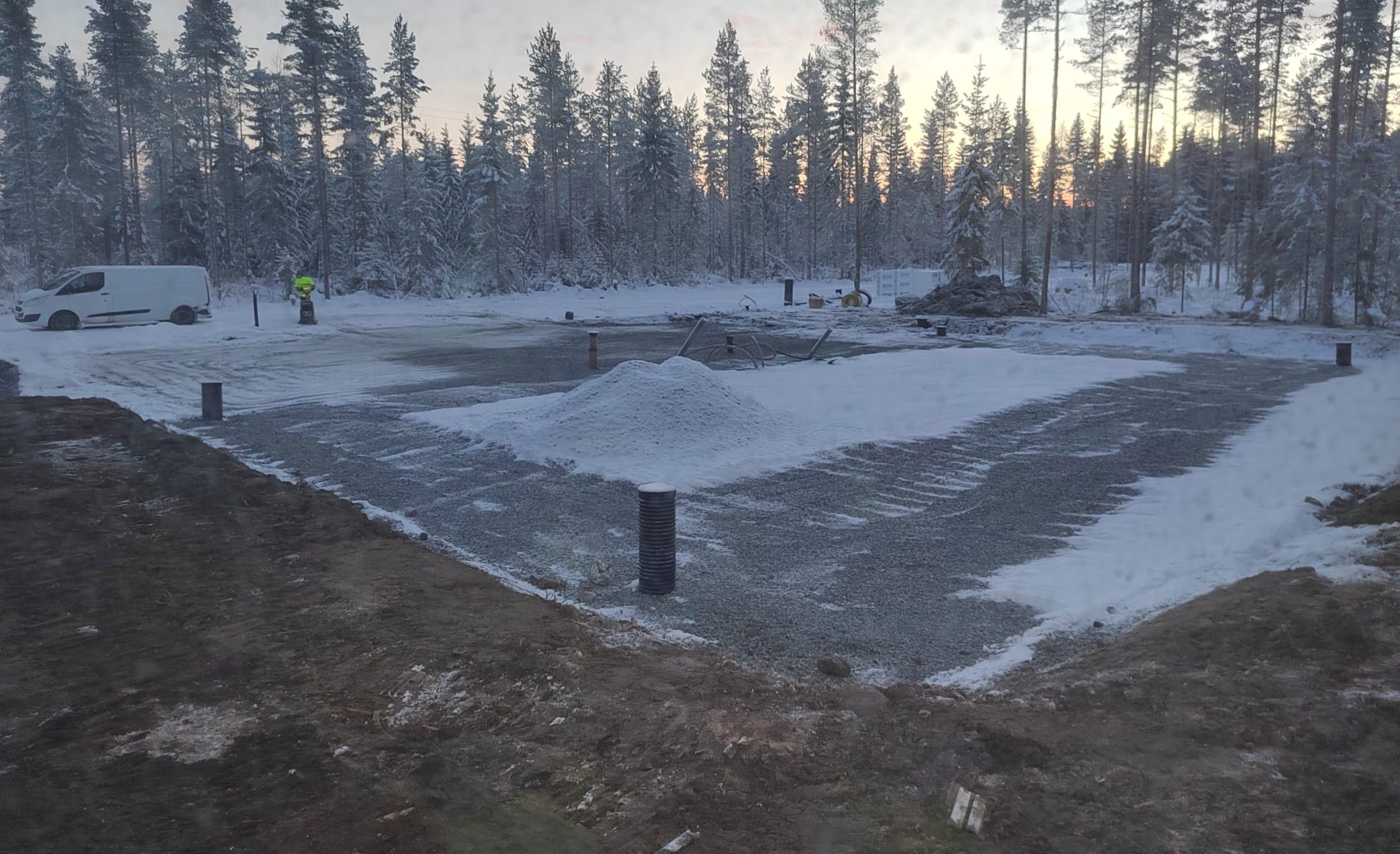 Talvinen maisema rakennustyömaalta, jossa näkyy lumipeitteinen pohja, kasa lunta keskellä, putkia ja työkoneita taustalla, sekä metsä ja auringonlasku horisontissa.
