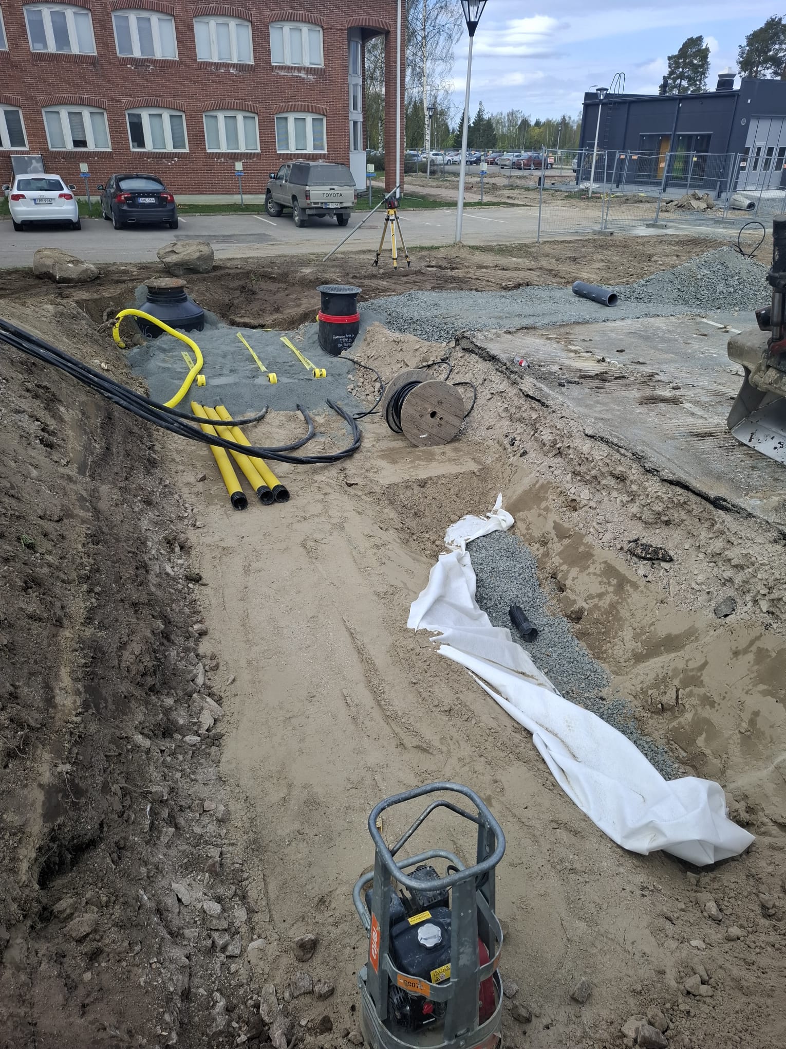 Rakennustyömaa, jossa on kaivantoja, putkia, kaapeleita ja tiivistyskone. Taustalla rakennuksia ja autoja. Valoisa sää.